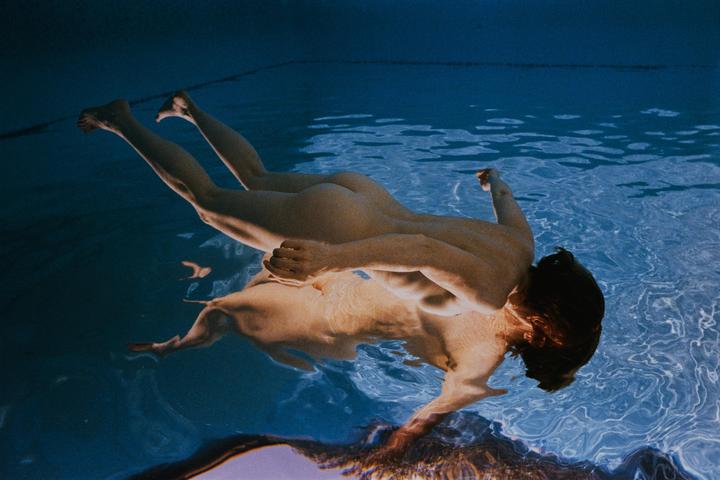 A nude man floats face down in a swimming pool, with his reflection visible on the water's surface.
