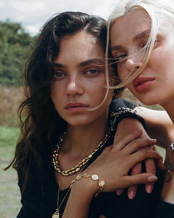 Two women wearing gold jewelry pose closely together, one with dark hair and the other with blonde hair.