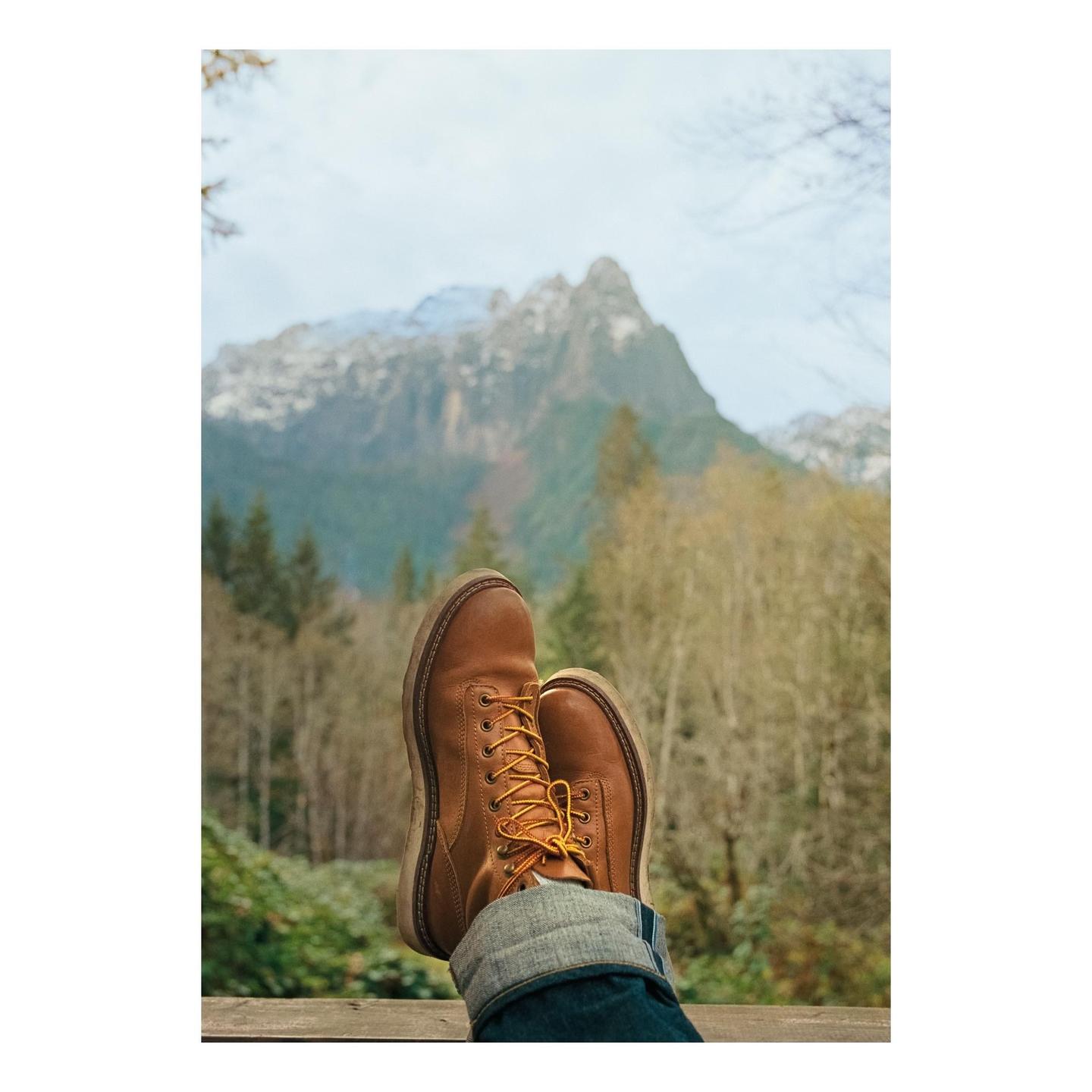 A person's feet in brown boots and cuffed jeans are propped up against a railing with a mountain and forest in the background.
