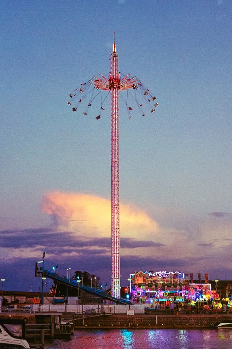 A tall swing ride is illuminated at a funfair next to a body of water at dusk.