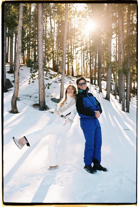 A couple in ski suits stands back-to-back in a snowy forest with sunlight streaming through the trees.