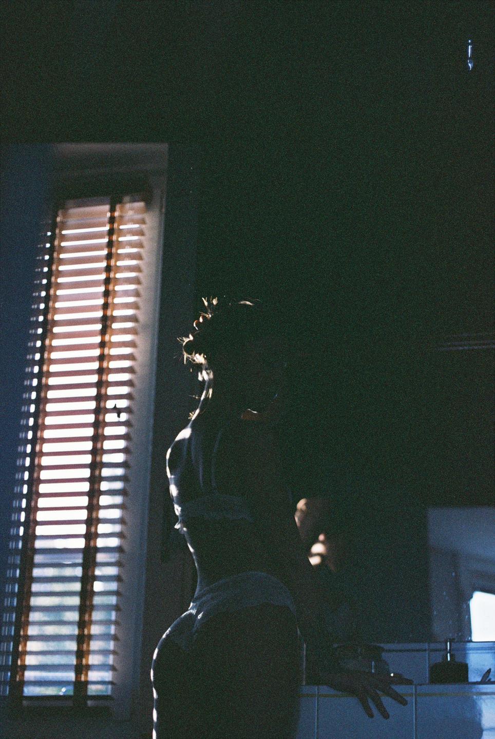 A woman stands in a dark room with light coming through a window with blinds.