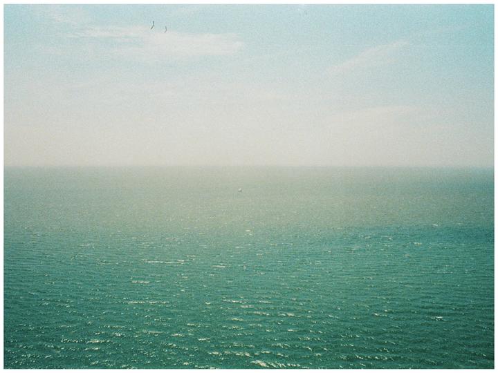 A vast expanse of ocean meets the sky on the horizon, with a small boat visible in the distance and two birds flying overhead.