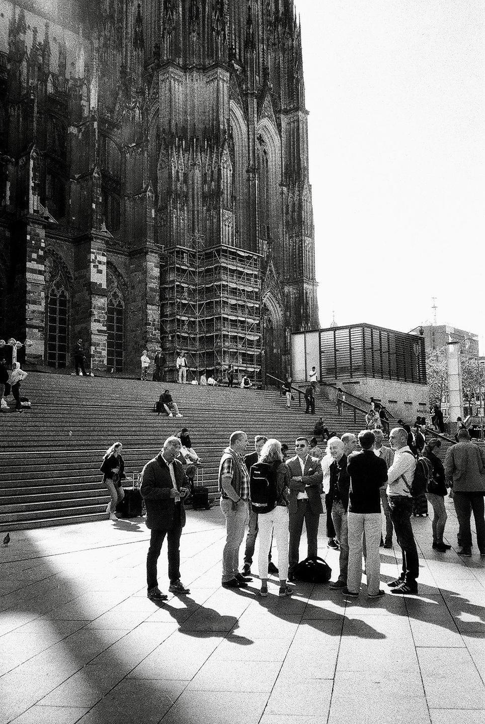 A large group of people are gathered on a paved area in front of a grand cathedral with scaffolding on its facade.
