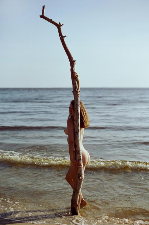 A nude woman stands in the ocean holding onto a large piece of driftwood.
