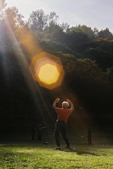 A woman practices tai chi in a park with a bright lens flare above her.