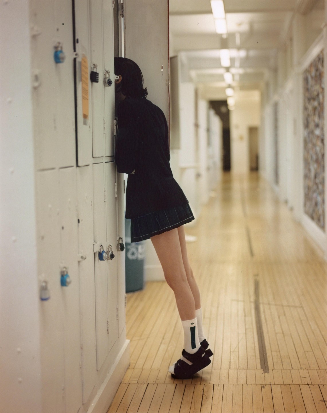 A girl in a plaid skirt and sandals leans against a locker in a school hallway.