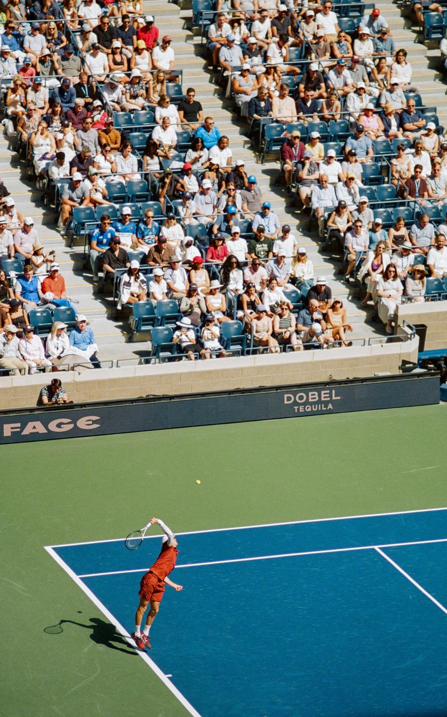 A tennis player serves on a blue and green court while spectators watch from the stands.