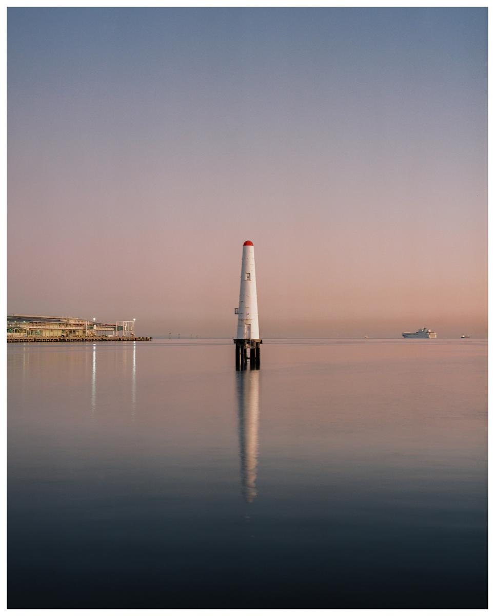 A white lighthouse with a red top stands in calm water with a building and a ship in the distance under a pastel sky.