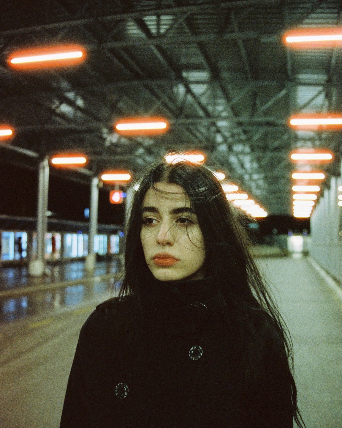 A woman with long dark hair stands on a train platform at night, looking to the side.