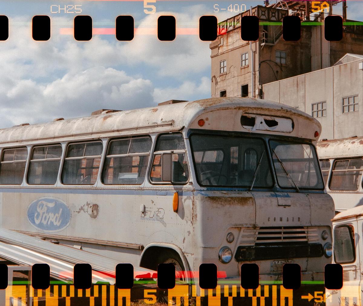 A weathered white bus with a Ford logo on its side is parked in front of a building under a cloudy sky.