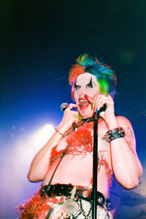 A person with colorful hair and clown makeup sings into a microphone on a stage.