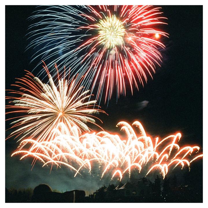 Fireworks explode in the night sky above a silhouetted crowd and trees.