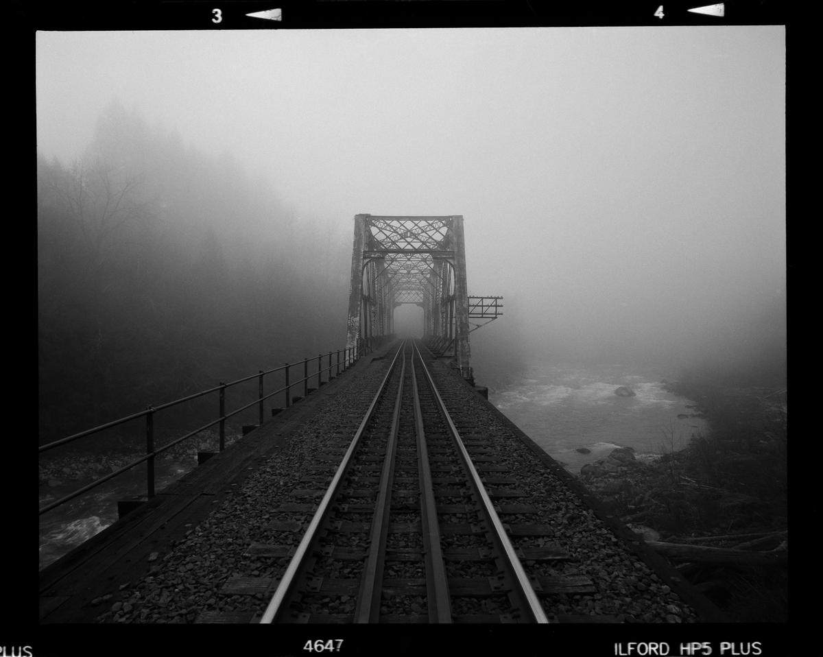 A train trestle extends into the fog over a river with trees on the left.