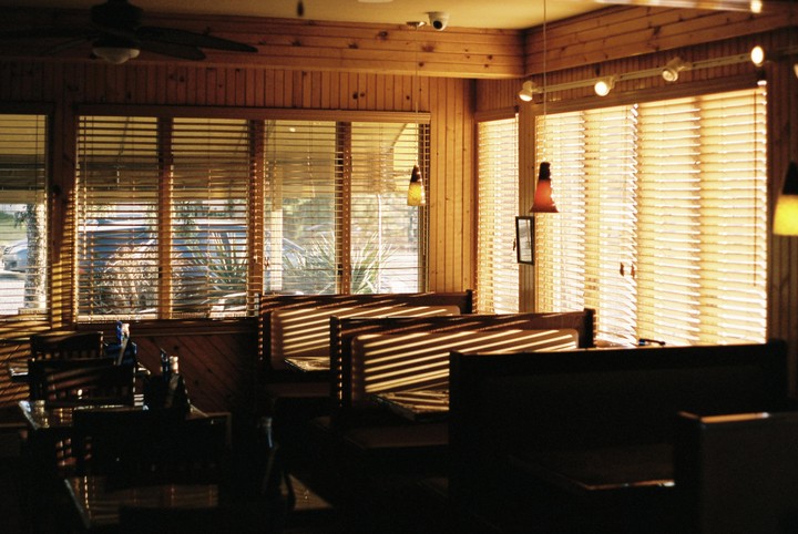 Sunlight streams through horizontal blinds, casting striped shadows across empty booths in a wood-paneled restaurant.