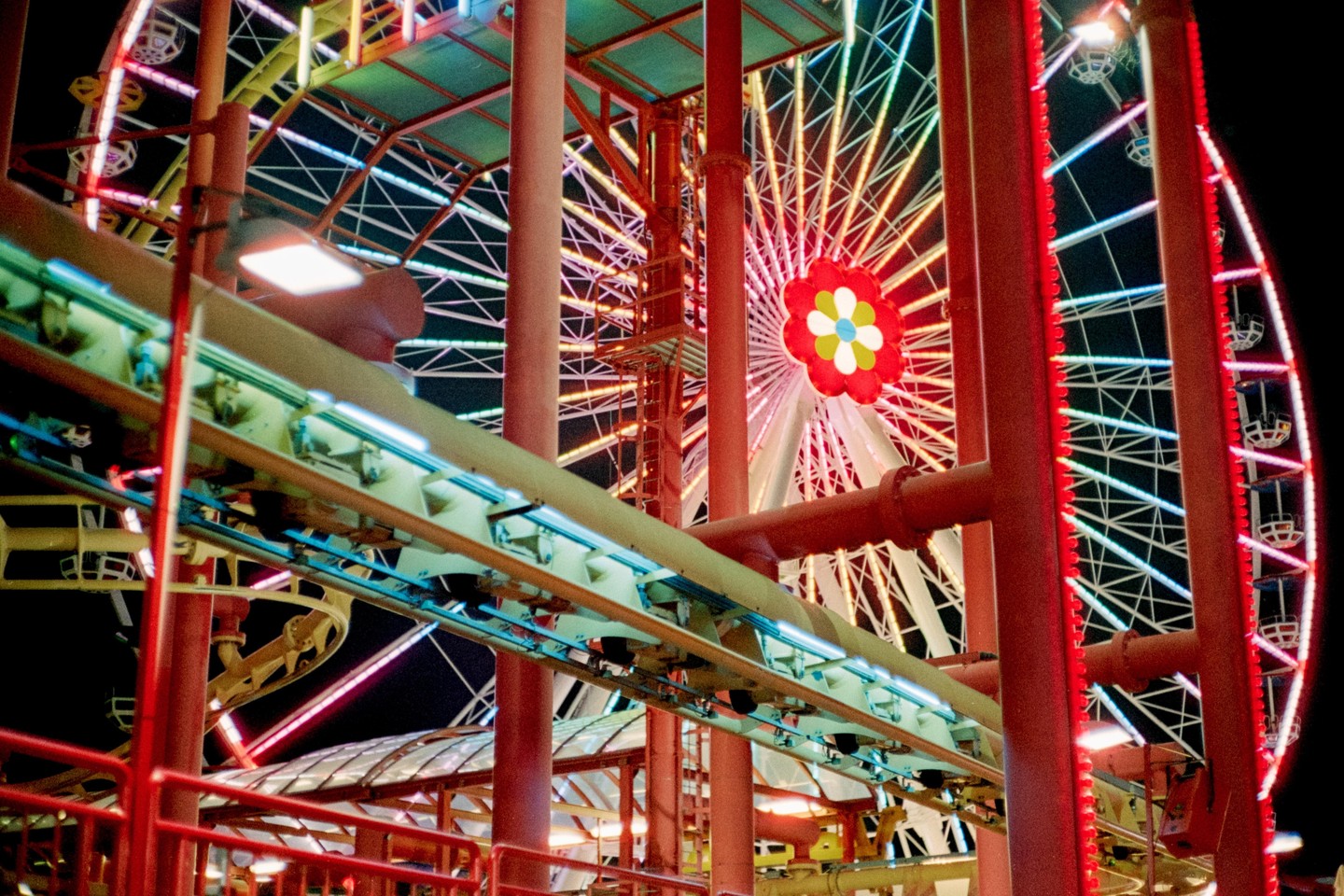 A brightly lit Ferris wheel with a flower design at its center is visible behind the tracks of a roller coaster at night.