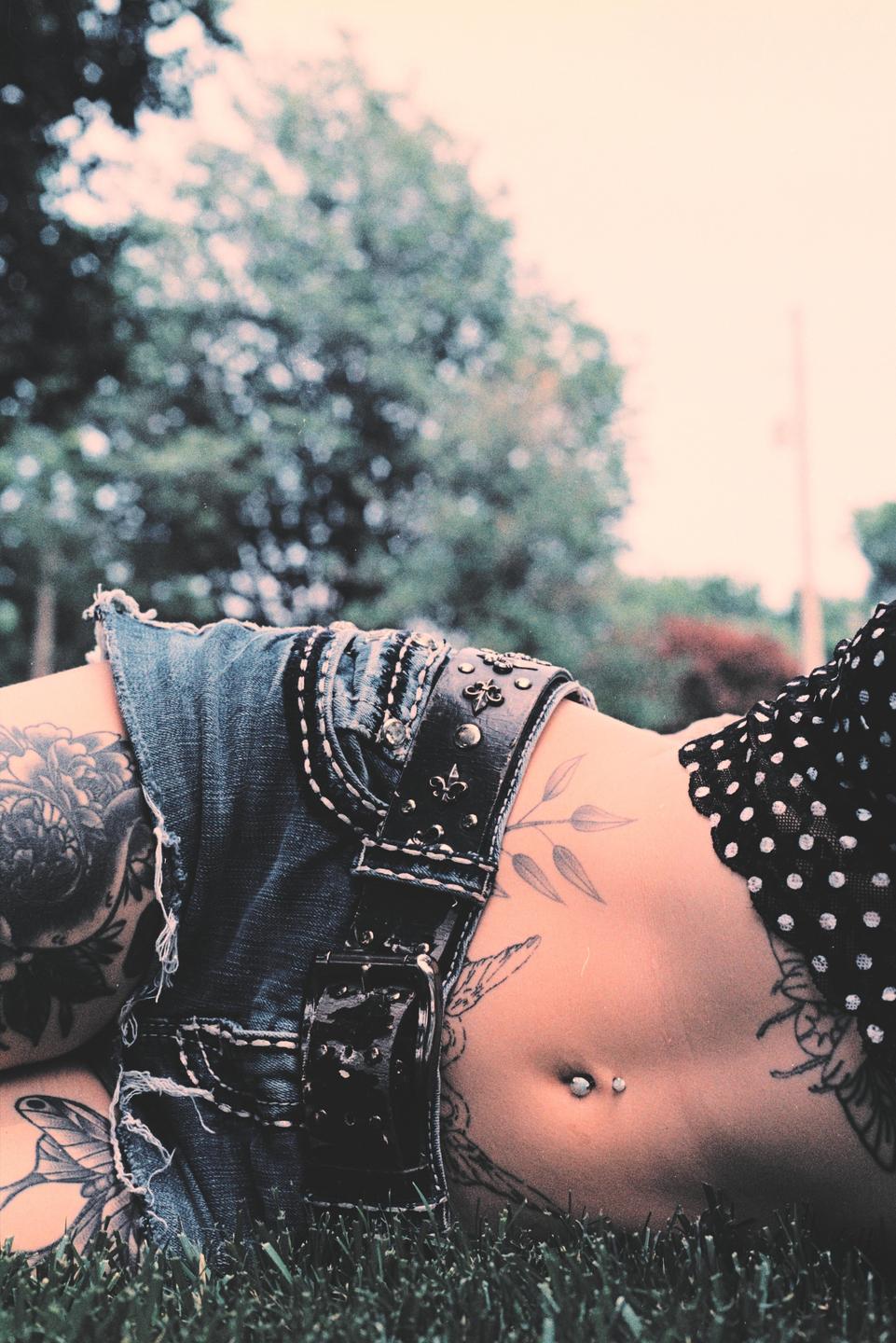 A woman with tattoos and a belly button piercing lies in the grass.