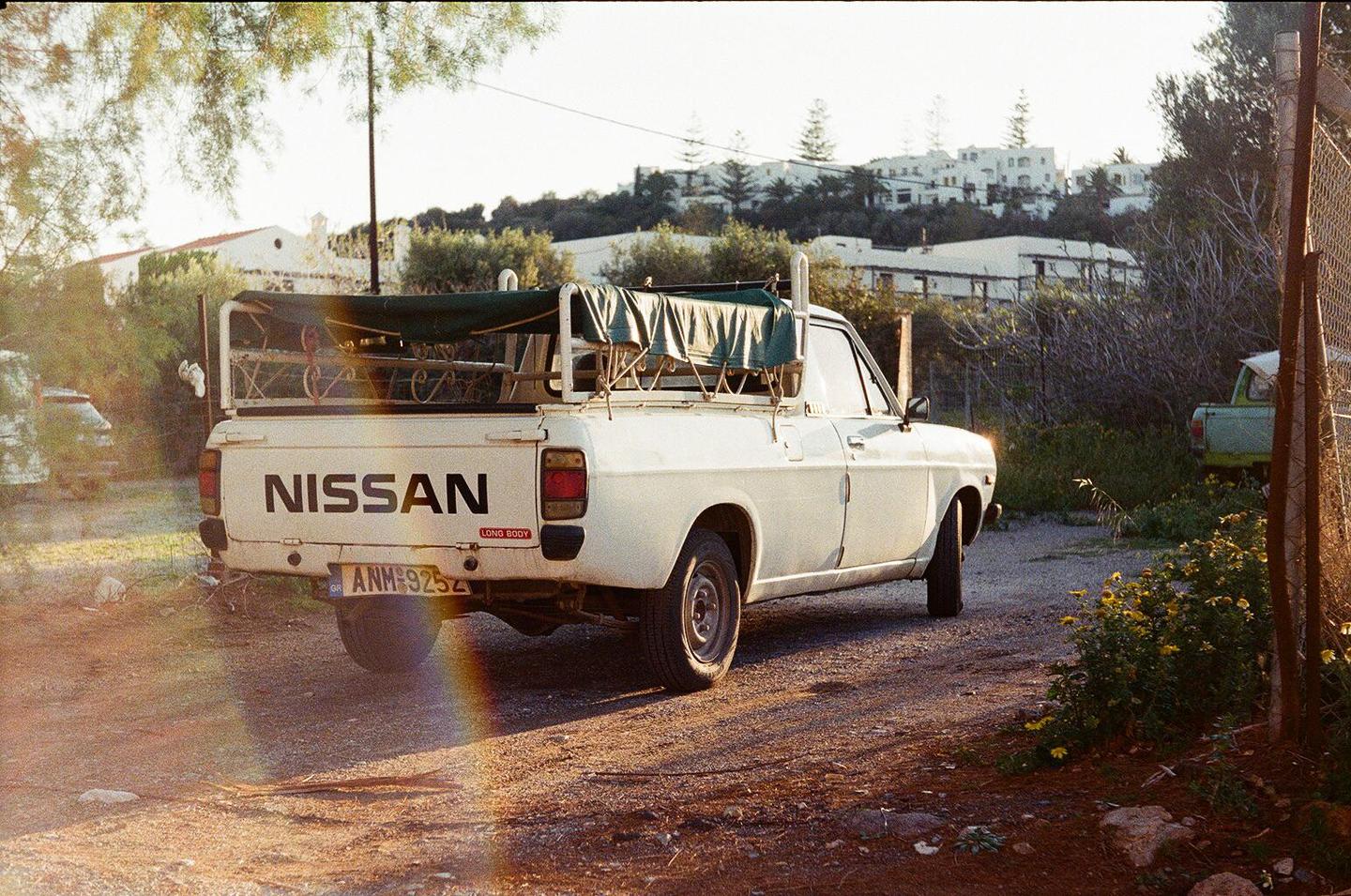 A white Nissan pickup truck is parked on a dirt road with a village on a hill in the background.