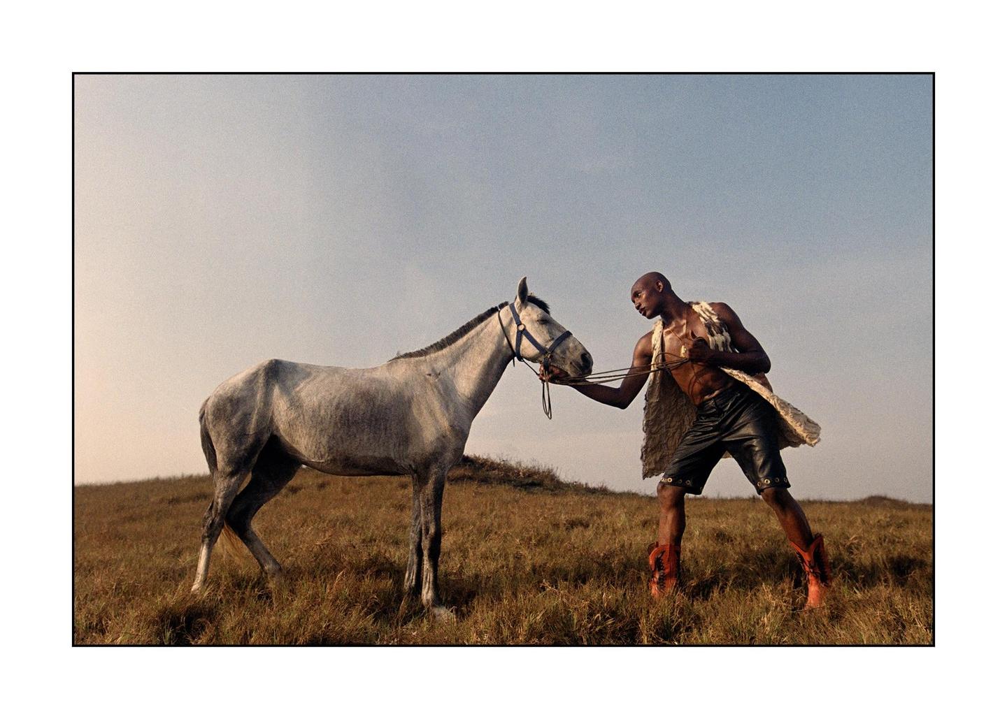 A man in a fur vest and cowboy boots holds the reins of a white horse in a field.