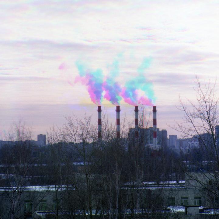 Four red and white striped smokestacks emit colorful smoke into a cloudy sky above a city.