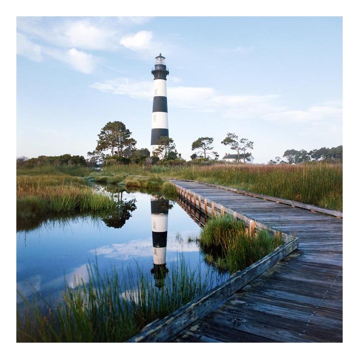 A black and white striped lighthouse is reflected in a still body of water next to a wooden boardwalk.