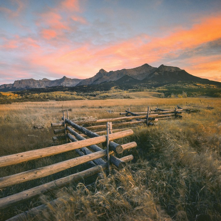 A wooden fence runs through a field of tall grass with mountains in the background under a colorful sky.