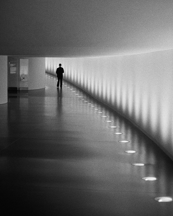 A man walks down a long, curved hallway with lights along the wall and floor.
