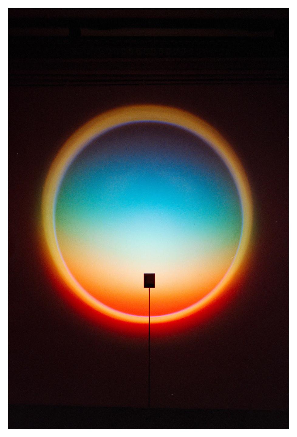 A circular light projection with a gradient of colors from blue to orange and red is cast onto a dark surface, with a small black square on a thin stand in the foreground.