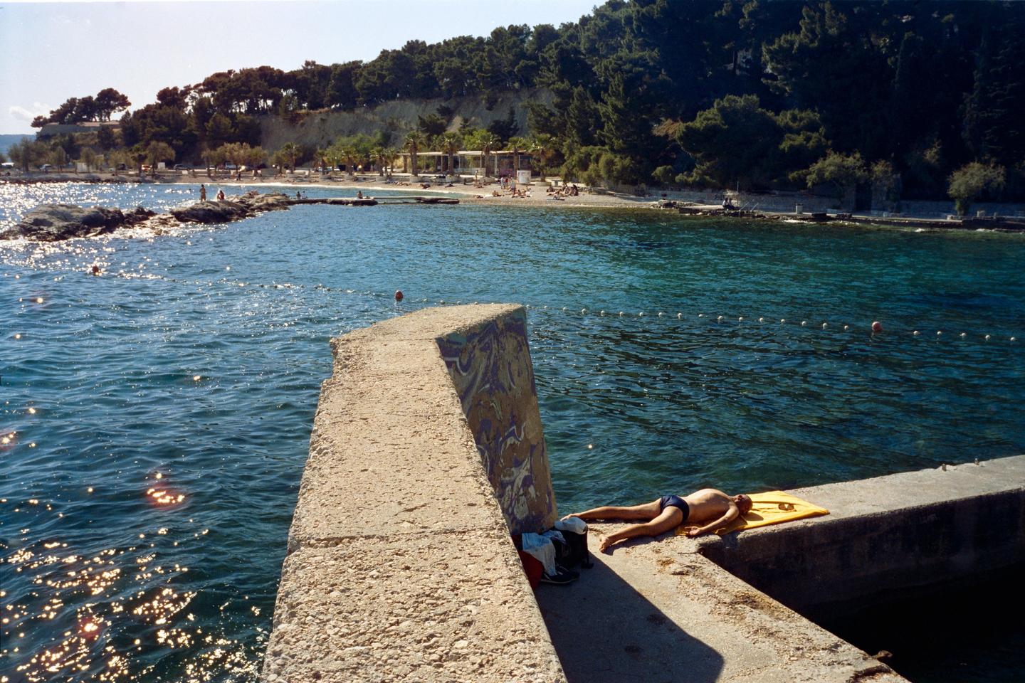 A man sunbathes on a concrete pier next to a body of water with a beach and trees in the distance.
