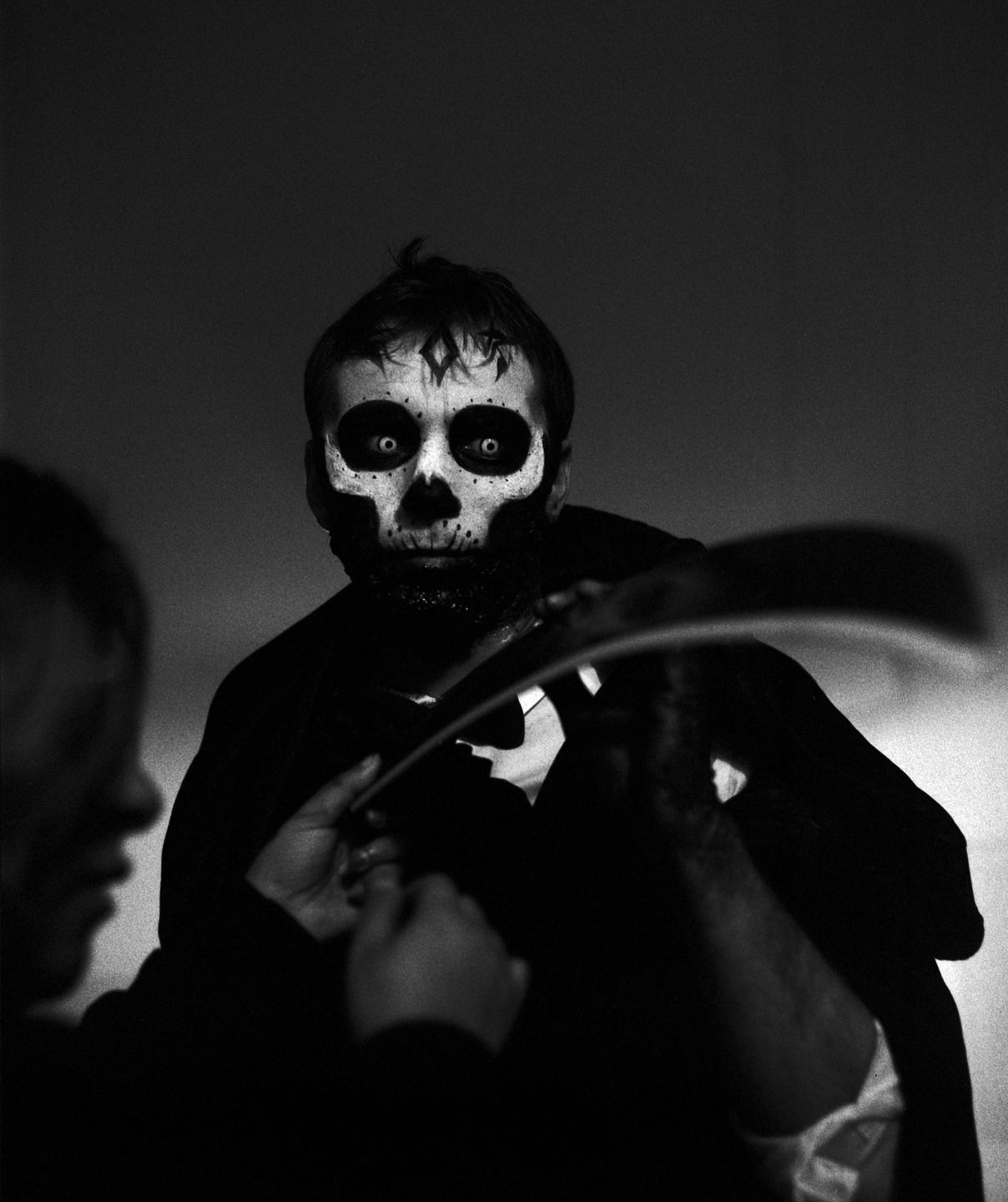 A man with skull makeup on his face looks directly at the viewer while another person's hands adjust his costume.