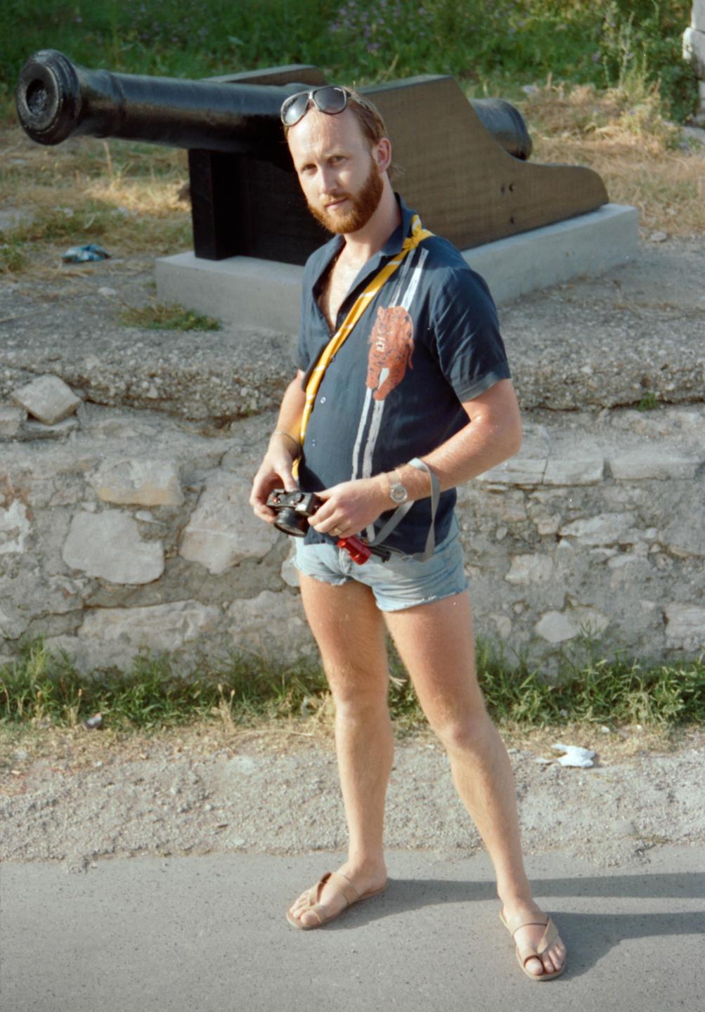 A man wearing sunglasses, a short-sleeved shirt, and denim shorts stands next to a cannon.