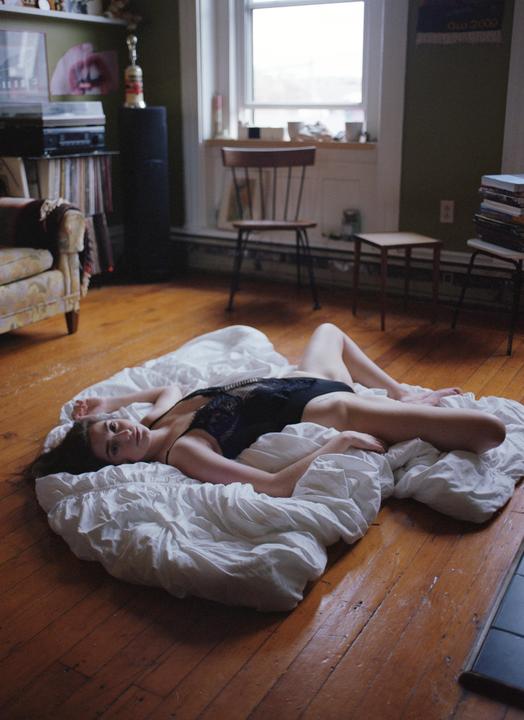 A woman in a black bodysuit is lying on a white sheet on a wooden floor in a room with a window.