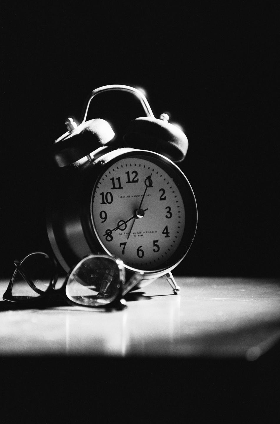 An alarm clock and a pair of spectacles are on a surface in a dark room.