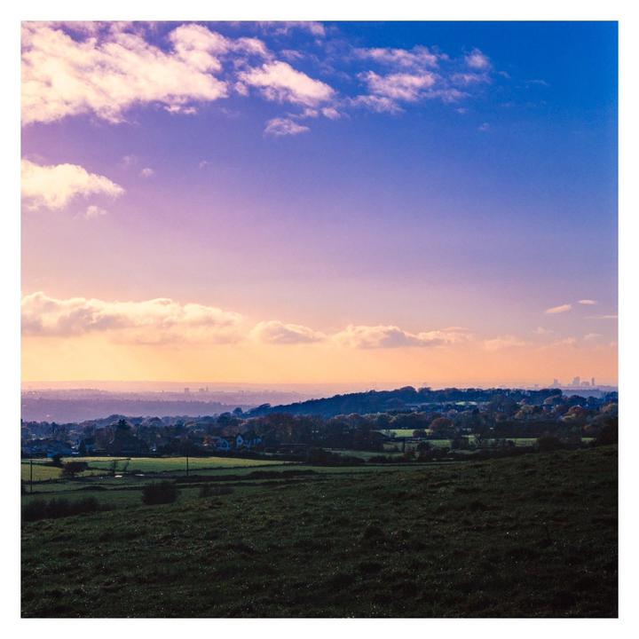 A wide landscape shows a field in the foreground, a town and hills in the middle ground, and a city skyline in the distance under a colorful sky.