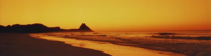 Waves crash on a beach with mountains in the background under an orange sky.
