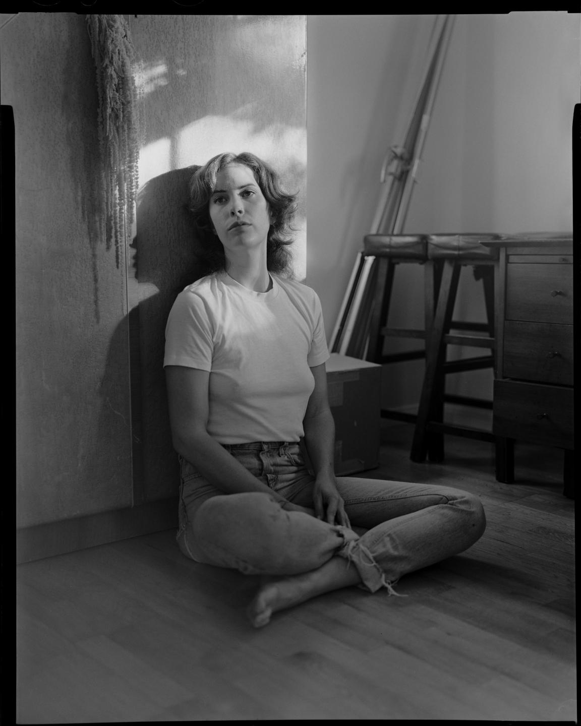 A woman sits on a wooden floor with her back against a wall, illuminated by a shaft of light.