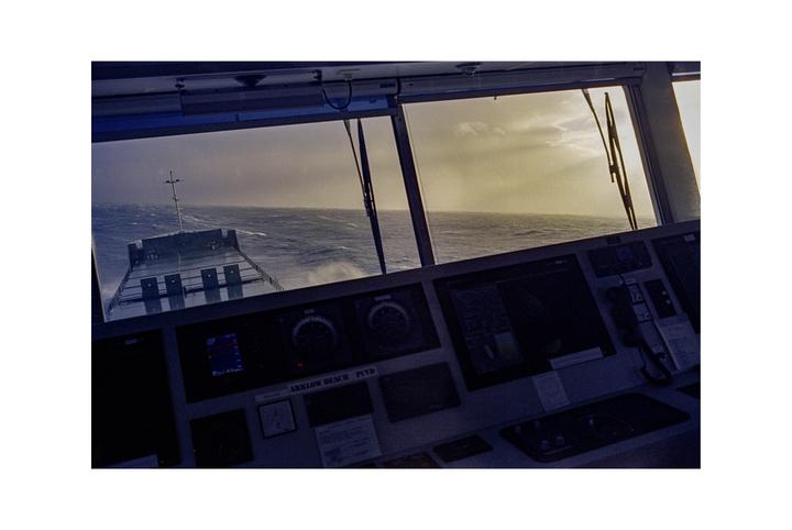 The view from a ship's bridge shows the deck of the ship and the rough sea under a cloudy sky.