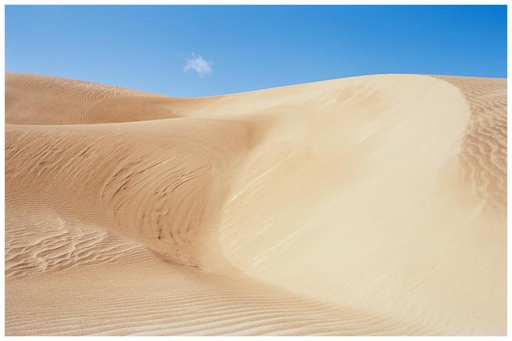 A single cloud floats above a desert landscape with sand dunes.