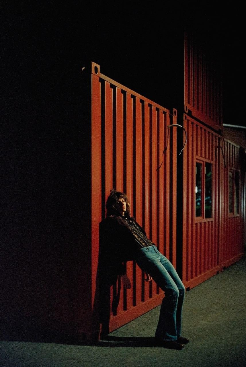 A woman leans against a red shipping container at night.