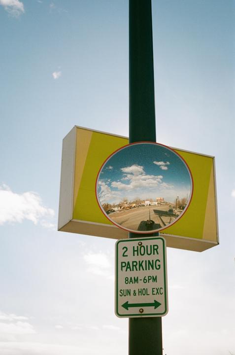 A convex mirror on a pole reflects a street scene and sky, with a parking sign below it.