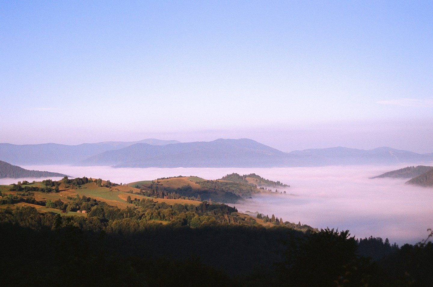A valley filled with fog is surrounded by hills and mountains under a clear sky.
