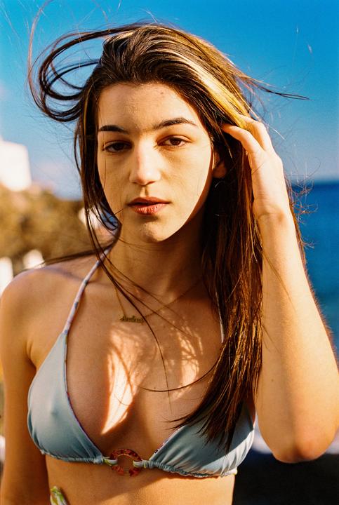 A woman with long hair and a bikini top looks directly at the viewer, with a bright blue sky and ocean in the background.