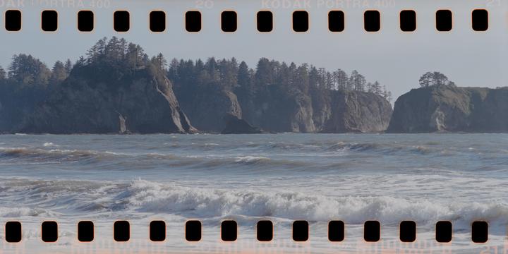 Waves crash on a beach with large, tree-covered cliffs in the background.