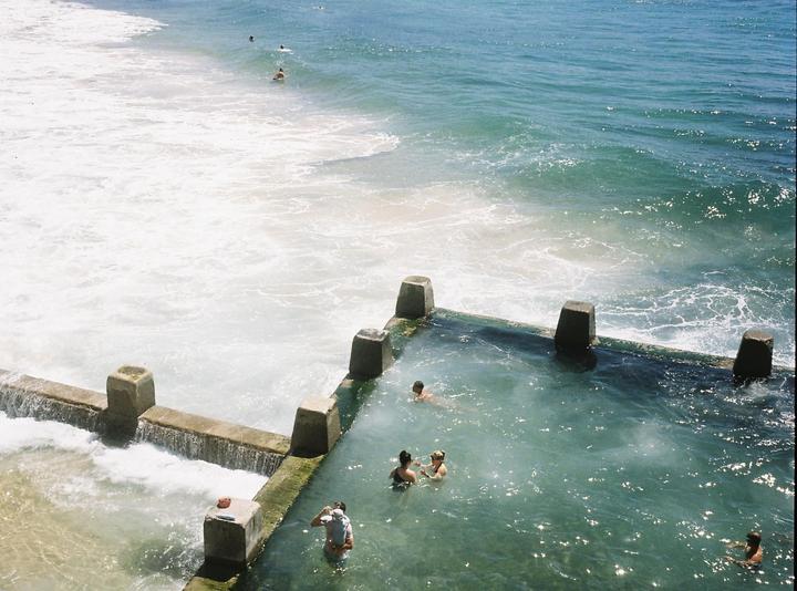 A concrete ocean pool with several people swimming in it is next to a foamy ocean.