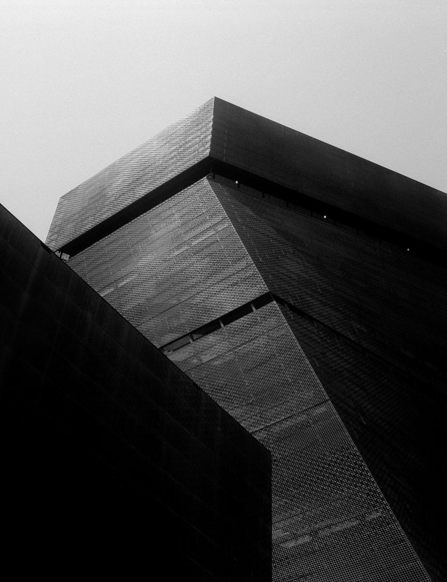 A low-angle view shows the geometric facade of the de Young Museum against a bright sky.