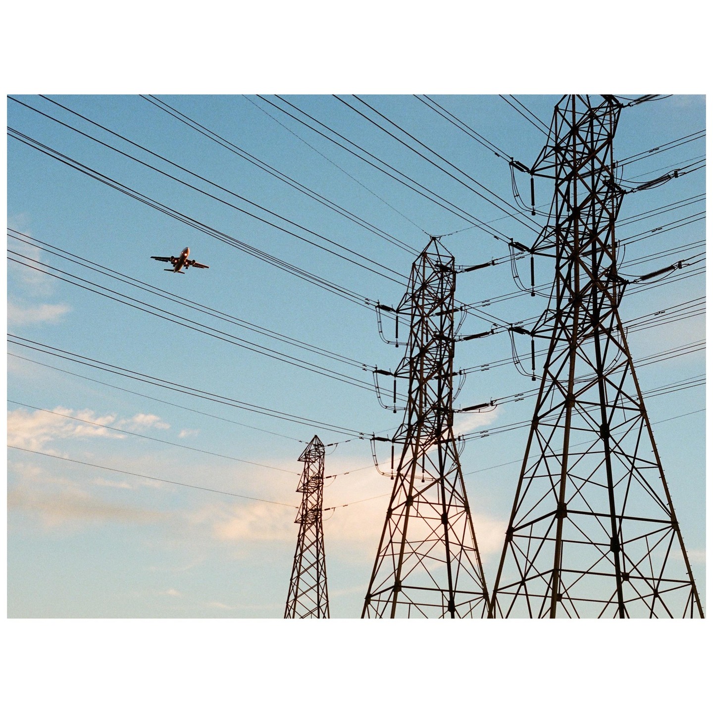 An airplane flies over three power line towers against a blue sky with scattered clouds.