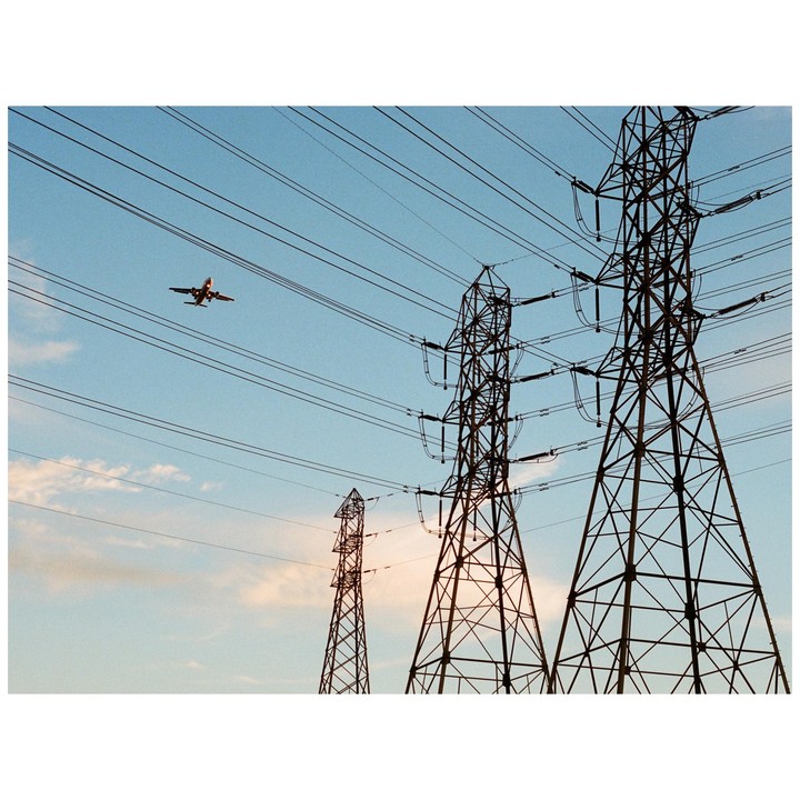 An airplane flies over three power line towers against a blue sky with scattered clouds.