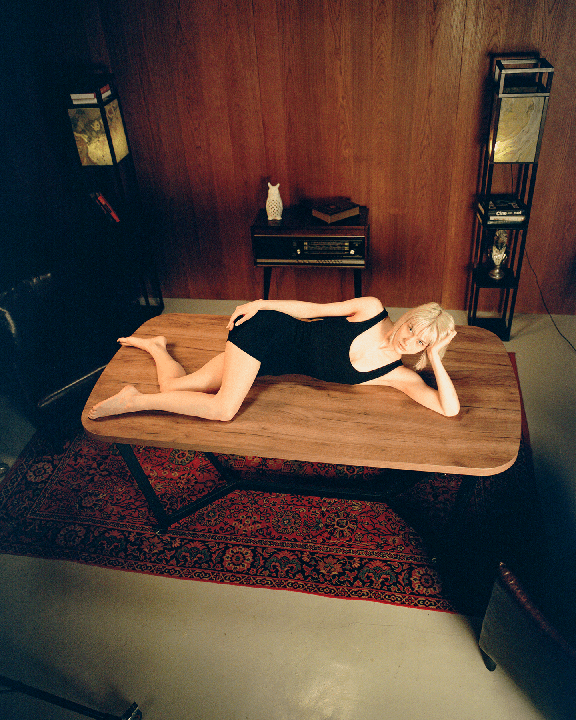 A woman in a black bodysuit is lying on a wooden table in a room with wood-paneled walls and a red patterned rug.