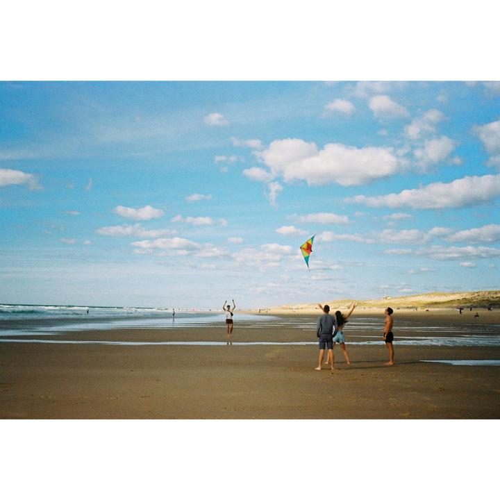 Three people stand on a beach looking at a kite flying in the sky while another person stands in the distance.