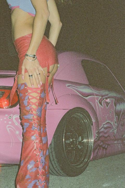 A woman in a red skirt and thigh-high boots stands next to a pink car.